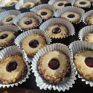 May include: Close-up of a tray of cookies. Each cookie is round with a light-colored center, a dark red filling, and a ring of chopped nuts around the edge. The cookies sit in white paper cups with gold foil rims.
