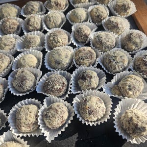 May include: Close-up of numerous chocolate truffles, each coated in a light-colored, textured layer. The truffles are nestled in individual white paper cups, arranged closely together. The dark chocolate centers contrast with the lighter coating.