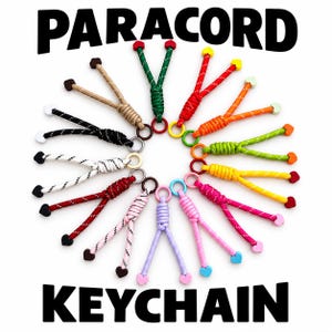 Op de afbeelding: Een verzameling paracord sleutelhangers gerangschikt in een cirkel, elk voorzien van een metalen ring, gevlochten koord en twee kleine hartvormige kralen aan de uiteinden. De sleutelhangers zijn verkrijgbaar in diverse kleuren, waaronder rood, groen, oranje, geel, roze, blauw, wit, zwart en beige.