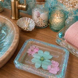May include: A clear, rectangular soap dish with a light blue glitter border, containing a light blue and pink pressed flower arrangement. The dish sits on a wooden surface, with a gold faucet and seashell accents in the background.