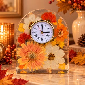 May include: An arched clear resin desk clock featuring embedded pressed flowers in shades of orange, yellow, and white. The clock face is white with black Roman numerals, a gold-tone bezel, and black hour and minute hands with a red second hand.