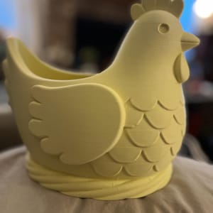 May include: A pale yellow chicken-shaped planter or decorative container. The object features a sculpted comb, a beak, a wattle, and layered wing and feather details. The base of the chicken is designed with a textured, rope-like pattern.