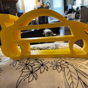 May include: A bright yellow, three-dimensional shelf shaped like the silhouette of a sports car with a rear spoiler. The structure features two horizontal levels for storage and circular cutouts representing the wheels of the vehicle.