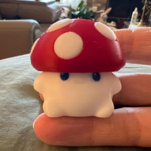 Puede incluir: Una figura con forma de hongo con un sombrero rojo y manchas blancas. La base es blanca con dos pequeños ojos azules. La figura se sostiene en la mano de una persona, mostrando su tamaño y diseño. Un artículo decorativo divertido.