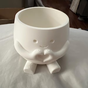 May include: A white ceramic planter with a whimsical design. The planter features a round body with a smiling face, small eyes, and arms forming a heart shape. It has two small feet at the bottom. The planter is designed for indoor or outdoor use.