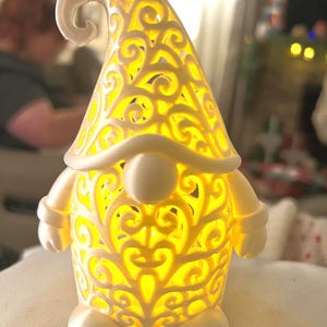 May include: A white gnome-shaped decorative lamp with a yellow glow emanating from within. The lamp features intricate, swirling cut-out patterns on its hat and body. The gnome has a round nose, arms, and feet.