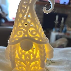 Peut inclure: Une figurine de gnome blanche et illuminée avec un motif ajouré détaillé. Le chapeau du gnome a une pointe recourbée et le corps brille d'une lumière chaude. La figurine a un visage rond, des bras et des pieds.