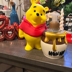 May include: A yellow Winnie the Pooh figurine wearing a red shirt stands next to a decorative soap dispenser. The dispenser is shaped like a honey pot with the word "HUNNY" on it. A red "Love" balloon is in the background.
