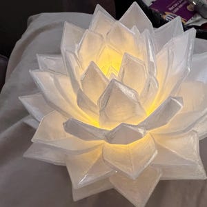 Pode incluir: Uma luminária de flor de lótus branca com pétalas translúcidas em camadas dispostas num padrão geométrico cristalino. O centro da flor emite um brilho amarelo quente e suave, iluminando a estrutura interna das pétalas por dentro.