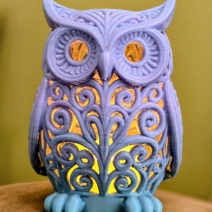 Puede incluir: Lámpara decorativa con forma de búho lavanda con intrincados patrones calados. El búho tiene ojos grandes y detallados y un diseño estilizado. La lámpara emite un brillo amarillo cálido, creando un ambiente suave. Los pies del búho son de un azul más claro.
