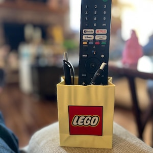 Può includere: Un contenitore giallo con il marchio LEGO che contiene un telecomando TV nero, una penna nera e una penna argentata. Il telecomando ha diversi pulsanti e i loghi di YouTube e Netflix. Il contenitore è appoggiato su una superficie beige.