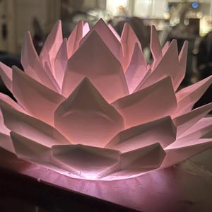 May include: A white, geometric lotus flower-shaped lamp. The lamp is lit from within, casting a soft pink glow. The petals are sharply pointed, creating a modern design. This decorative lamp is suitable for home decor.