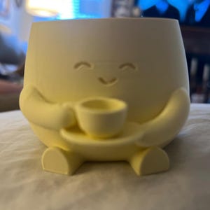 May include: A light yellow ceramic planter shaped like a smiling character, holding a small teacup and saucer. The planter has rounded features and a cheerful expression, perfect for small plants or succulents.
