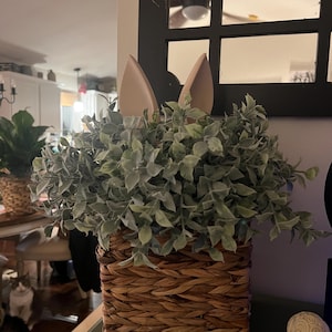 May include: A woven basket filled with faux greenery and adorned with light brown bunny ears. The basket has a rectangular shape and is made of natural-colored woven material. The greenery is a mix of green and gray leaves.