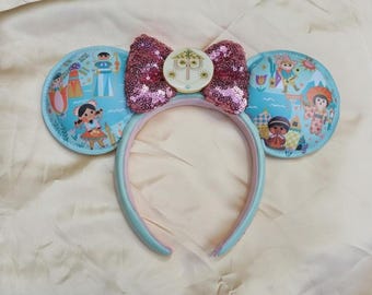 May include: A headband featuring two light blue circular ears decorated with colourful illustrations of various characters and landmarks. A large, shimmering pink sequined bow sits in the centre, accented by a circular white emblem with a stylised face.
