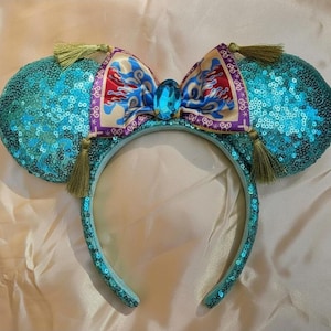 Può includere: Fascia per capelli con orecchie di topo turchese con paillettes con un fiocco fantasia con accenti rossi, blu e oro. L'arco è centrato con una grande gemma blu. Le orecchie hanno nappe dorate.