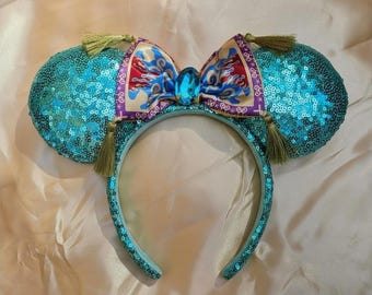 May include: Turquoise sequined mouse ear headband with a patterned bow featuring red, blue, and gold accents. The bow is centered with a large blue gem. The ears have gold tassels.