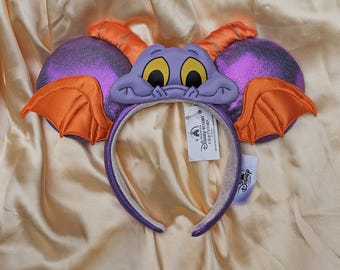 May include: A character ears headband in purple and orange. The ears are purple with orange wings and accents, and the headband is purple and white. The character has yellow eyes and a purple face. A Disney Resort tag is attached.