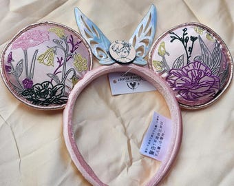 May include: Pink floral embroidered mouse ears headband with a light blue fairy wing accent and a small circular image. The ears are round with embroidered flowers. The headband has a tag with text.