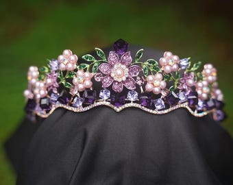May include: A decorative crown with a floral design. The crown features purple and pink flowers, green leaves, and sparkling crystals. The base is adorned with square-cut purple gems and a scalloped edge.