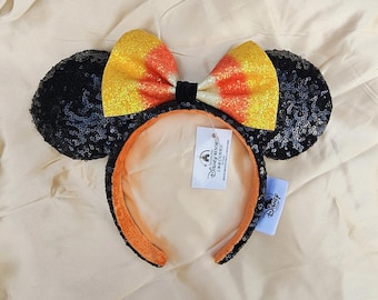 May include: A Halloween-themed headband with black sequined mouse ears and a candy corn-coloured bow. The headband has an orange interior and a black exterior. A Disney Resort tag is attached.