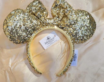 May include: Gold sequined Minnie Mouse ear headband with a matching bow. The headband has a gold band and is covered in small, shiny sequins. A Disney Resort tag is attached.