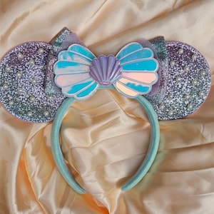 May include: A headband featuring two circular ears covered in purple and silver glitter with small white pearls attached. A central bow is made of iridescent blue material shaped like a seashell with a lavender shell accent in the center.