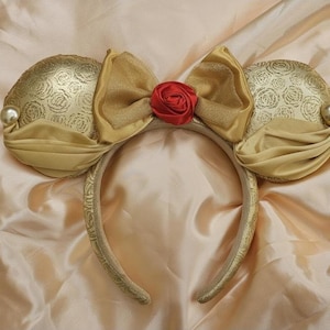 Könnte beinhalten: Goldfarbenes Mausohren-Stirnband mit einer großen Schleife und einer roten Rose in der Mitte. Die Ohren haben ein Rosenmuster und sind mit einer Perle verziert. Das Stirnband ist goldfarben mit einem dekorativen Muster.