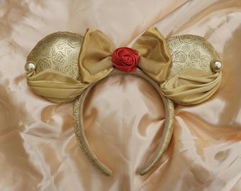 May include: Gold-coloured mouse ear headband featuring a large bow and a red rose. The ears have a rose pattern and are decorated with a pearl. The headband is gold with a decorative pattern.