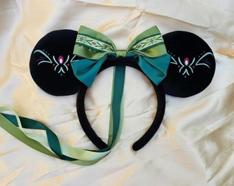 May include: Black mouse ear headband featuring a teal and green bow and ribbons. The black ears are decorated with a teal, pink, and white design. The headband is displayed on a cream-colored surface.