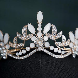 Pode incluir: Uma tiara dourada com detalhes de pedras preciosas brancas e strass transparentes. A coroa apresenta um design vertical central e motivos florais e de folhas. A tiara está sobre uma superfície preta.
