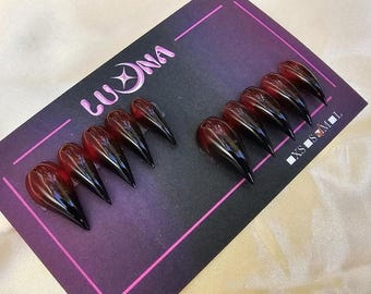May include: A set of ten long, pointed, press-on nails with a gradient effect. The nails transition from a deep red at the base to black at the tips. They are displayed on a dark purple card with the brand name "Luona" in pink.