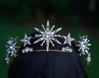 May include: A gold-coloured crown with star and crystal embellishments. The crown has a central starburst design and smaller star clusters. The tiara is set against a dark background, with a gold-coloured band.