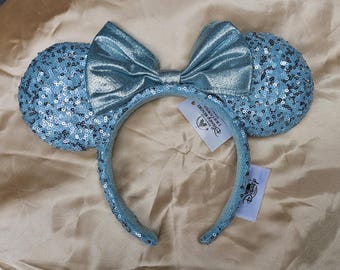May include: A light blue sequined headband with matching ears and a large bow. The headband has a Disney Resort tag attached. The ears and headband are covered in small, shiny sequins. The bow is made of a shimmering, light blue fabric.