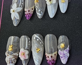 May include: A collection of ten almond-shaped false nails. Each nail has a grey base with diverse designs, including white swirls, floral accents, and gold leaf details. Some nails feature 3D flower embellishments and purple accents.