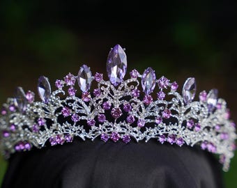 May include: A silver tiara with an intricate, decorative pattern. The crown is embellished with several purple gemstones of different sizes, including large, oval-shaped stones and smaller, round ones. The tiara is set against a dark background.