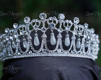 May include: A silver tiara adorned with clear rhinestones and pear-shaped faux pearls. The crown has an elaborate design with arches and dangling pearl accents. The tiara is displayed on a black surface against a blurred green backdrop.