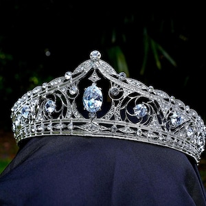 Könnte beinhalten: Eine silberne Tiara mit aufwendigem Filigran und zahlreichen klaren, funkelnden Edelsteinen. Die Krone hat einen großen, oval geformten Mittelstein und ist vor dunklem Hintergrund platziert.