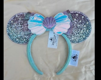 May include: A headband with two round, glittery ears in shades of blue and purple. A bow in the centre features a seashell design with blue, pink, and purple accents. The headband is light blue. Includes a Disney tag.