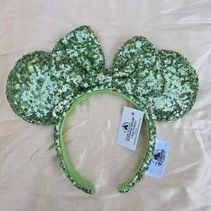 Puede incluir: Diadema de orejas de ratón con lentejuelas verdes y un lazo a juego. La diadema y las orejas están cubiertas de lentejuelas verdes brillantes. La diadema tiene una etiqueta de Disney Resort.
