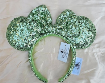 May include: Green sequined mouse ear headband with a matching bow. The headband and ears are covered in shimmering green sequins. The headband has a Disney Resort tag attached.