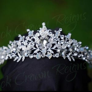 Puede incluir: Una tiara plateada con cristales y gemas transparentes. La tiara presenta un diseño floral y de hojas, con una gema central más grande. El fondo es verde borroso, con el texto "Black Crown Reigns".