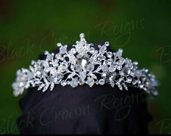 May include: A silver tiara adorned with clear crystals and gemstones. The design incorporates floral and leaf motifs, with a prominent central gemstone. The background is a blurred green, with the text "Black Crown Reigns" visible.