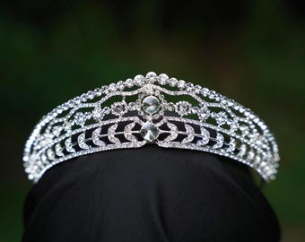 May include: A silver tiara with a detailed, openwork pattern. It is adorned with many clear, sparkling crystals and a prominent central crystal. The design incorporates leaf-shaped elements, giving it an elegant and decorative look.