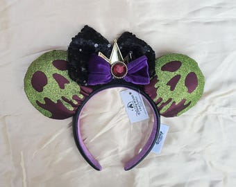 May include: A black sequined bow and purple velvet bow with a gold crown and red jewel accent sit atop a black headband. Green glittery ears with a skull design and a Disney Resort tag complete the design.