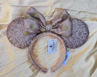 May include: A rose gold sequin headband featuring two circular ears and a large central bow with gold polka dots. The headband includes a white tag with the Disney Resort logo and text. The sequins cover the entire surface of the ears and band.