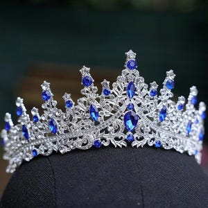 May include: A silver crown featuring detailed filigree work, set with numerous oval and round sapphire-blue gemstones. The crown is displayed on a black surface, highlighting its ornate design.