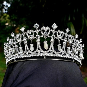 May include: A silver tiara with clear crystals and dangling faux pearls. The crown features a heart-shaped design and is set against a dark background, with a blurred green backdrop.