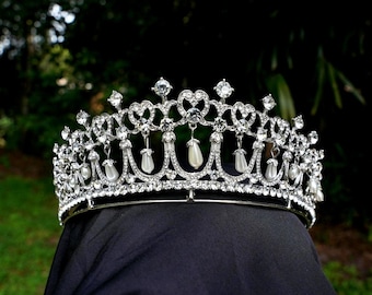 May include: A silver tiara adorned with clear crystals and faux pearl drops. The crown has a heart-shaped pattern and is set against a dark background, with a blurred green backdrop.