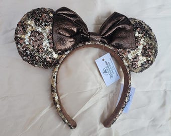 May include: A brown sequined Minnie Mouse ear headband with a matching bow. The ears are covered in a mix of brown, gold, and silver sequins. The bow is a solid, shiny brown. A Disney Resort tag hangs from the headband.
